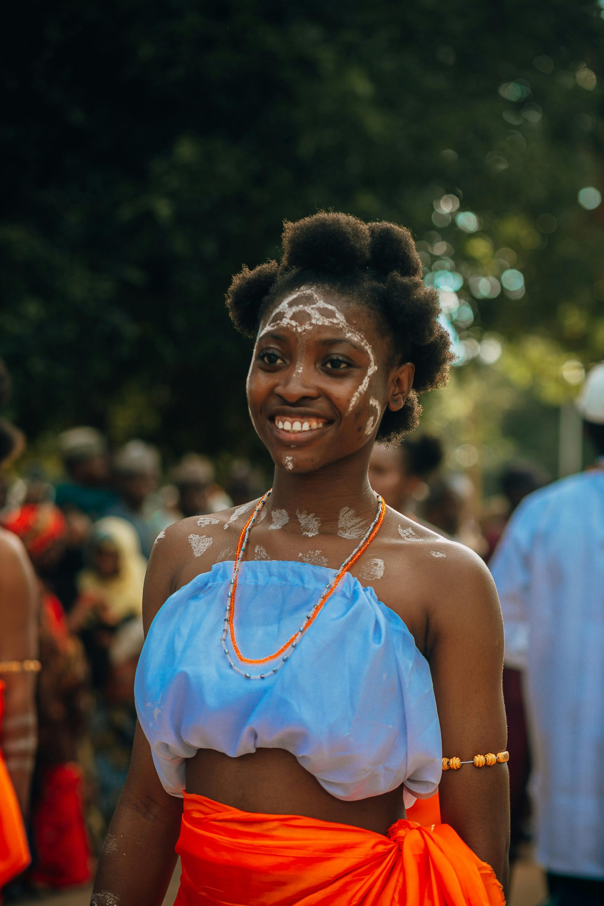 Young woman in vibrant traditional attire with cultural face paint at a lively outdoor festival.